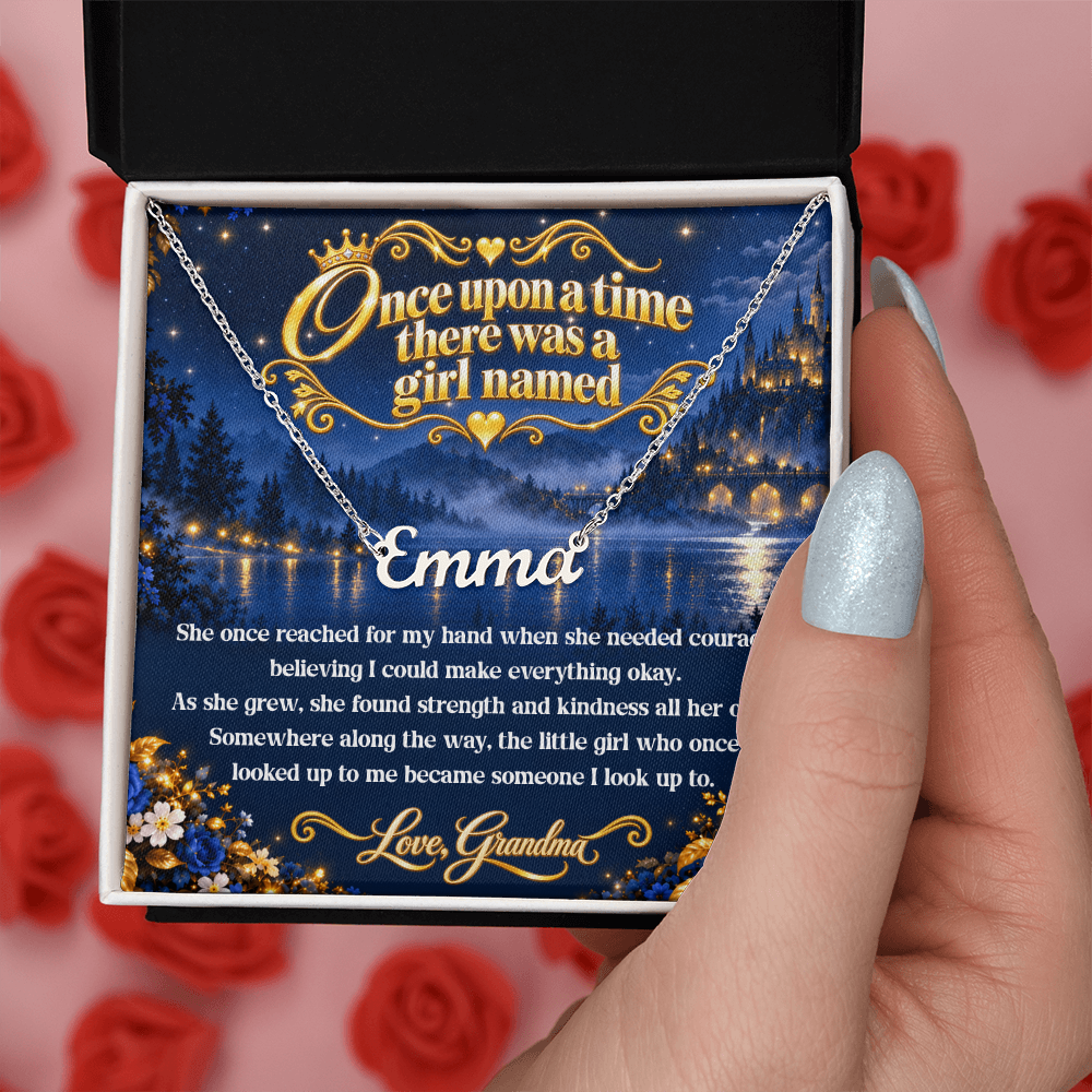 Hand holding a silver-tone Emma name necklace and blue fairy-tale card from Grandma in an open black and white gift box.