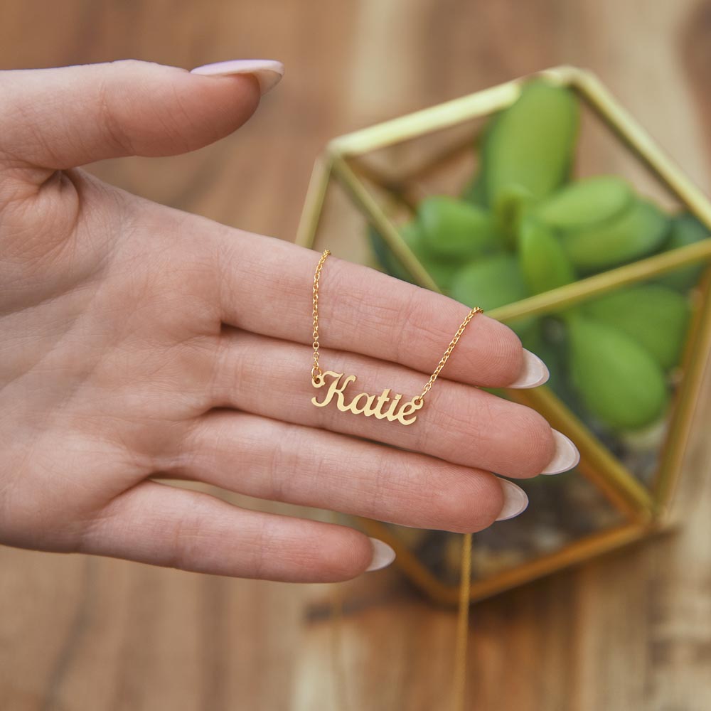 Gold-tone Katie name necklace draped across a hand beside a glass planter.