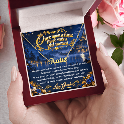 Hands holding a wood-style gift box with a gold-tone Katie name necklace and fairy-tale card from Grandma, beside pink roses.